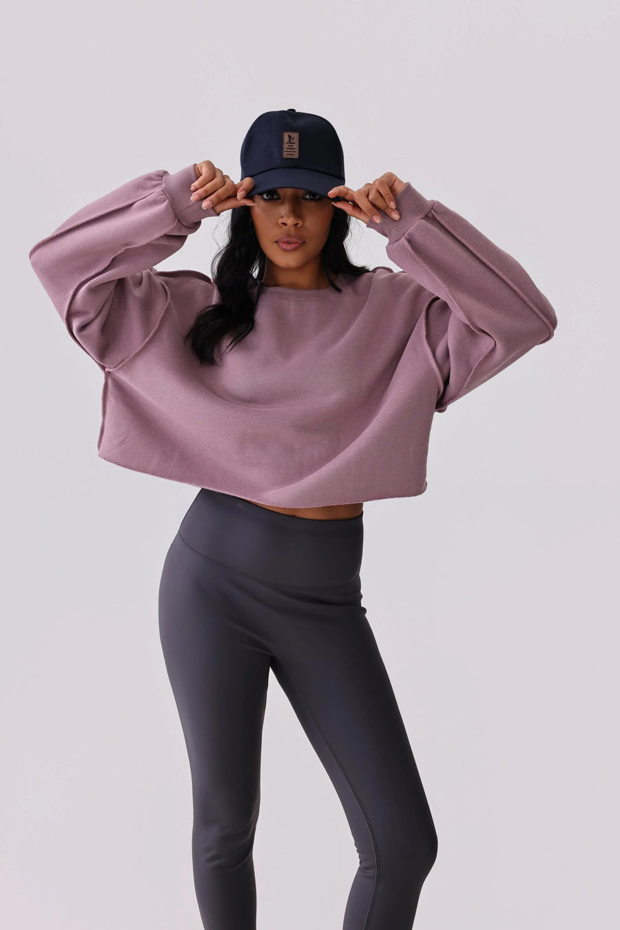Woman in mauve cropped sweatshirt and gray leggings, adjusting navy cap, activewear outfit