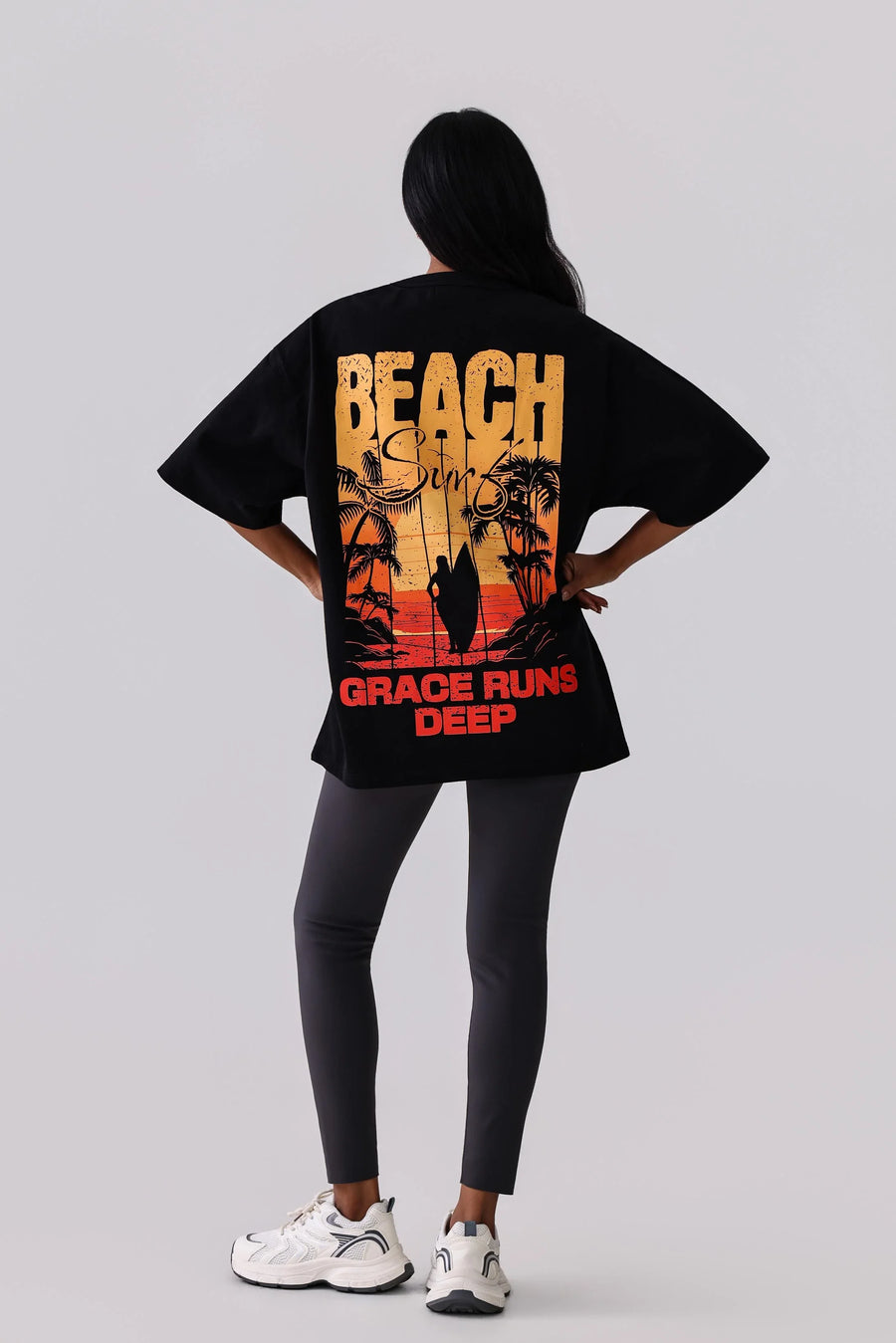 Black graphic tee with beach surf design, palm trees, and 'Grace Runs Deep' text, paired with leggings and sneakers.