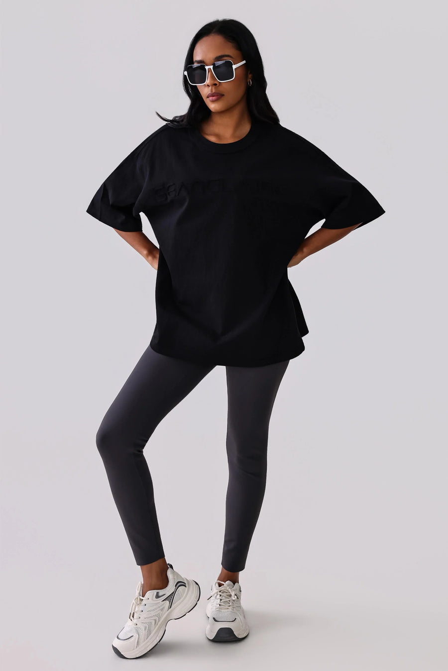 Woman in black oversized activewear tee, gray leggings, and white sneakers, styled with sunglasses for gym or streetwear.