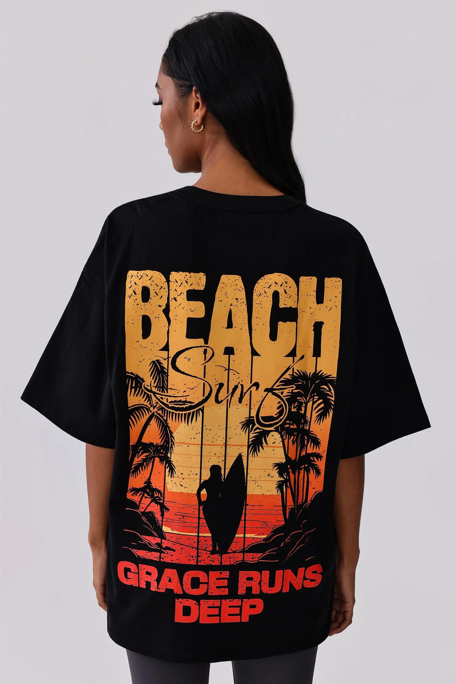 Black oversized graphic tee with beach surf sunset print, palm trees, surfer silhouette, and 'Grace Runs Deep' text